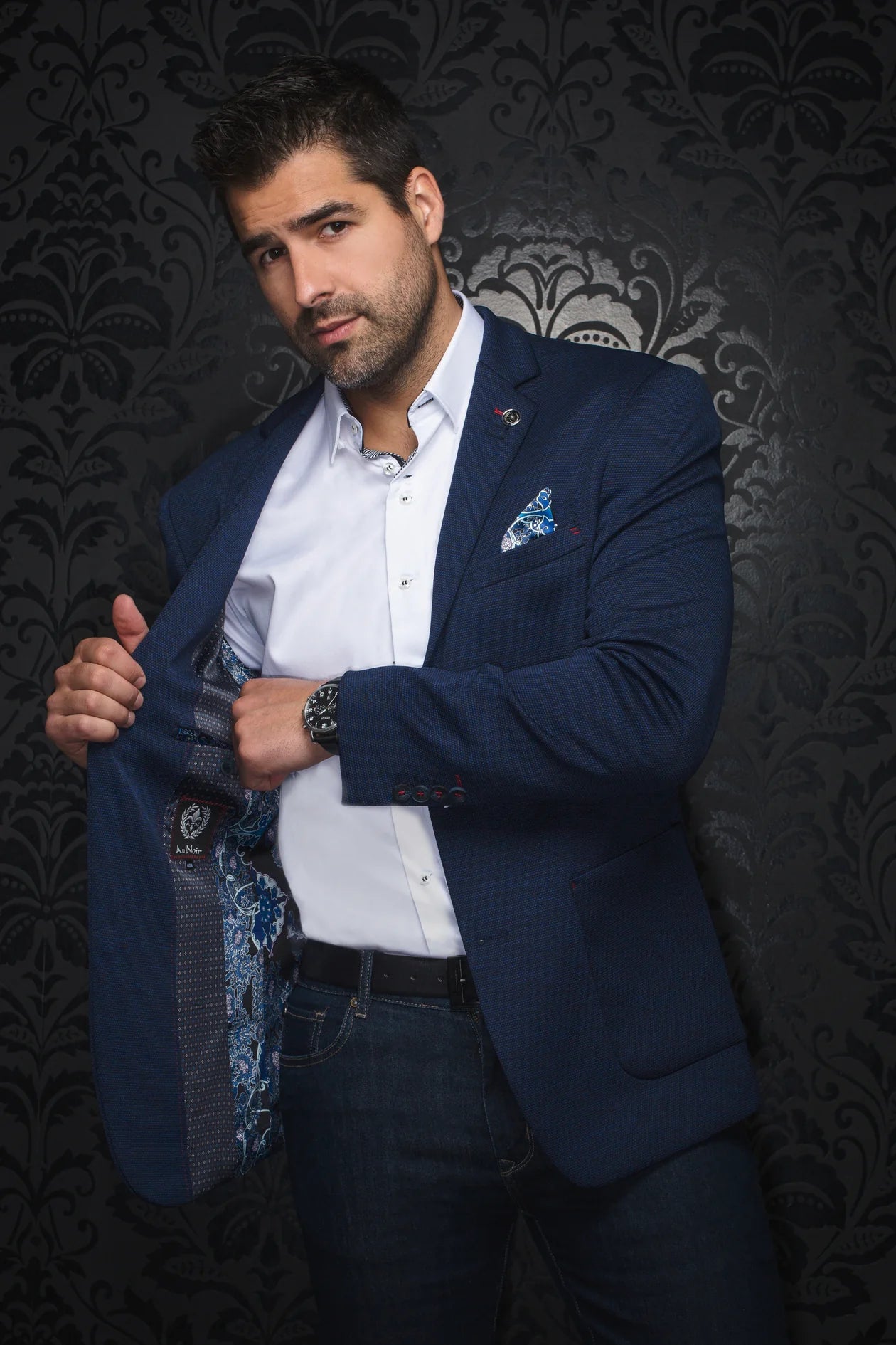 Man wearing a blue blazer over a white shirt with a patterned tie against a dark, decorative wall.