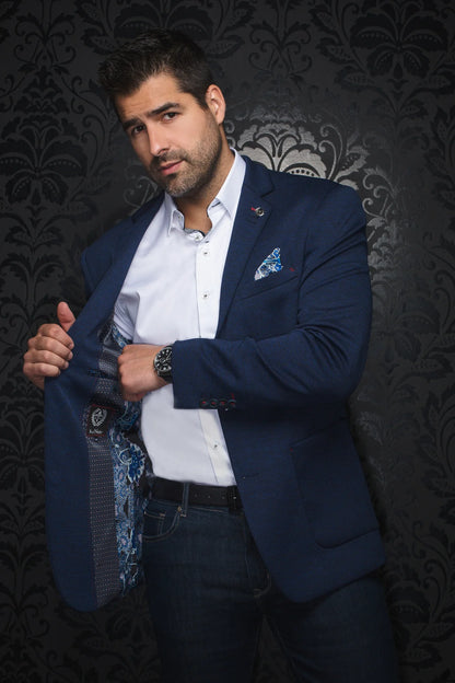 Man wearing a blue blazer over a white shirt with a patterned tie against a dark, decorative wall.