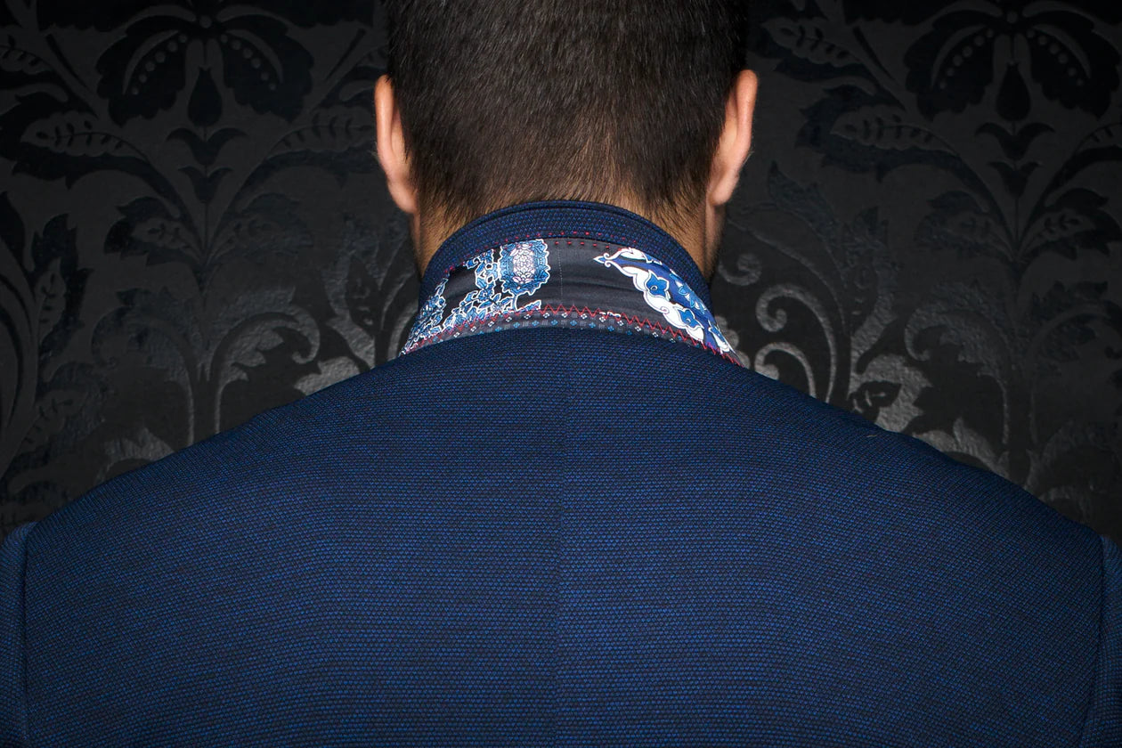 Back view of a person wearing a blue suit with a patterned shirt collar against a dark, decorative wall.