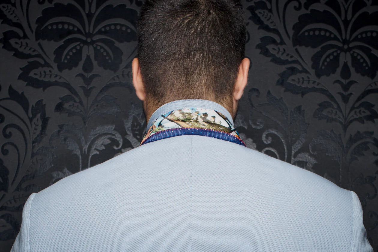 Person wearing a light gray suit with a patterned tie against a dark, decorative wall.