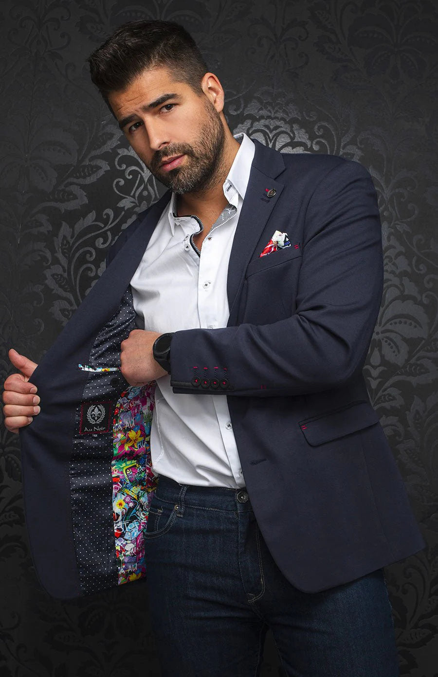 Man wearing a dark blue blazer with a colorful tie over his shoulder against a dark patterned background