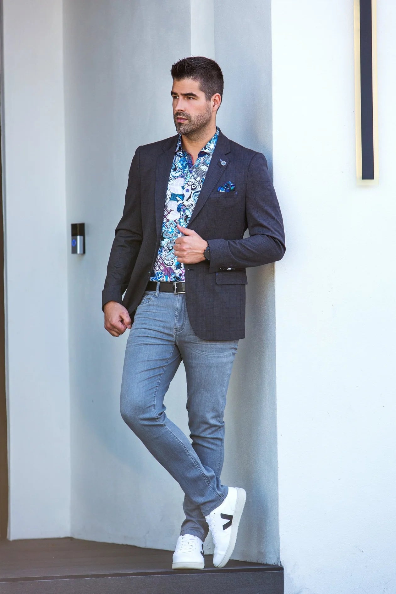 Man wearing a dark blazer, floral shirt, light jeans, and white sneakers in an indoor setting.