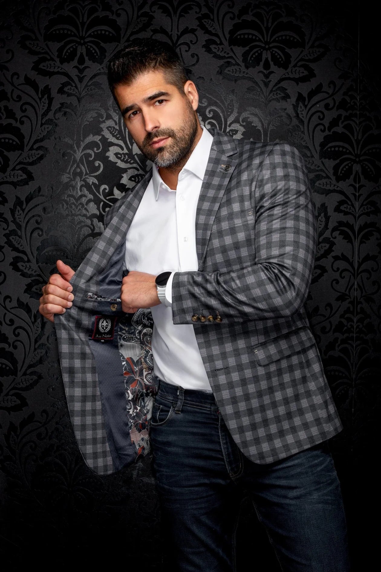Man wearing a checkered blazer over a white shirt with a patterned tie against a dark, decorative background.