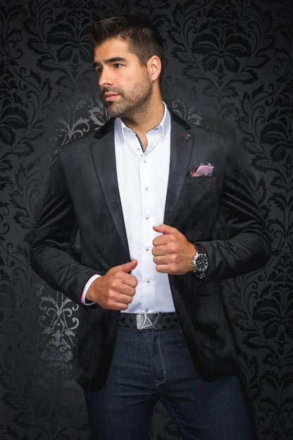 Man wearing a black blazer over a white shirt with a patterned pocket square, standing against a dark, ornate wall.