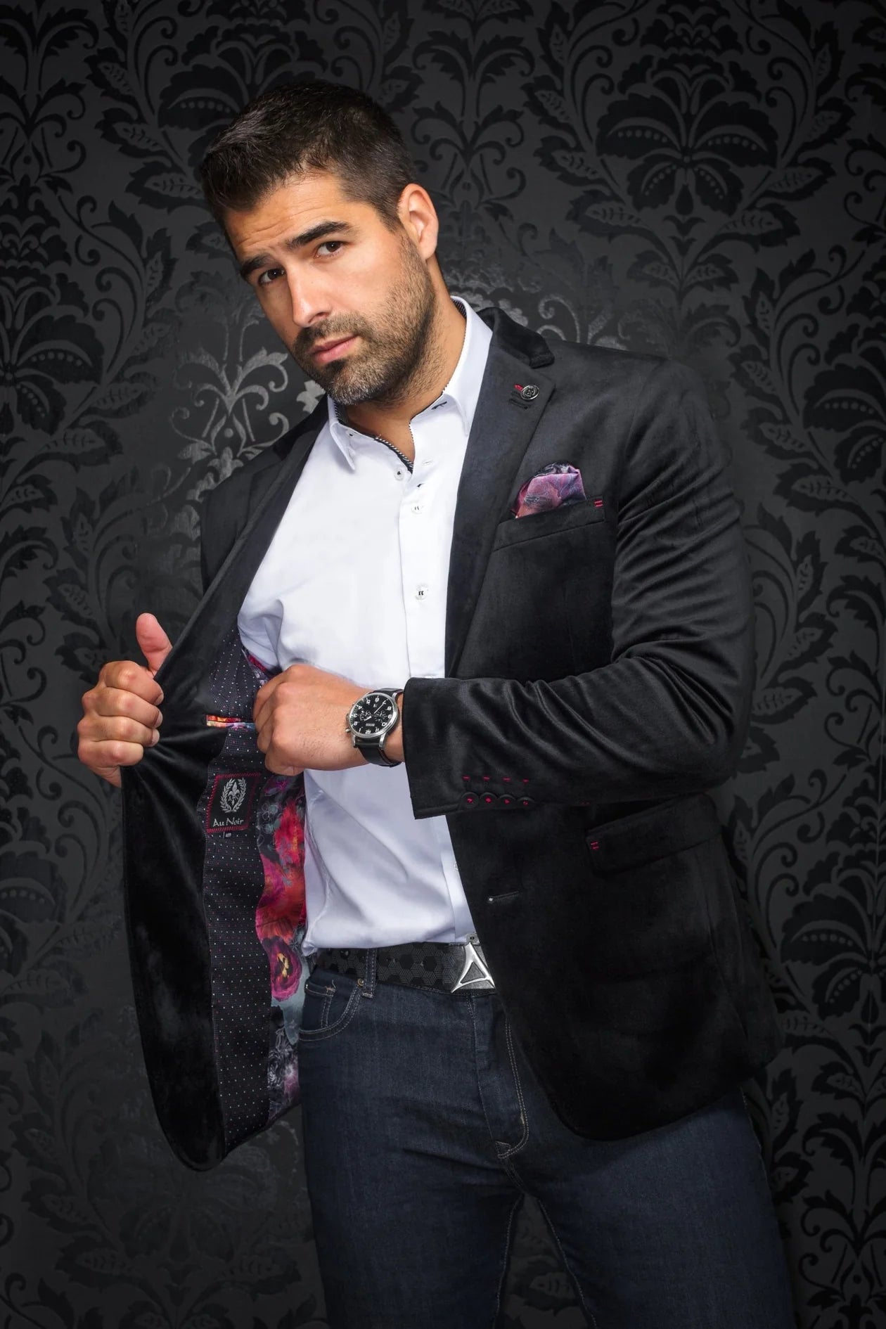 Man in a black blazer with a patterned pocket square against a dark, decorative background
