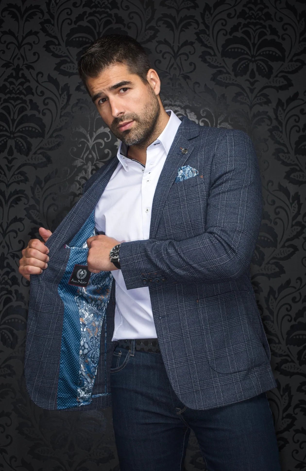 Man wearing a blue checkered blazer over a white shirt with a floral pocket square, against a dark patterned wall.
