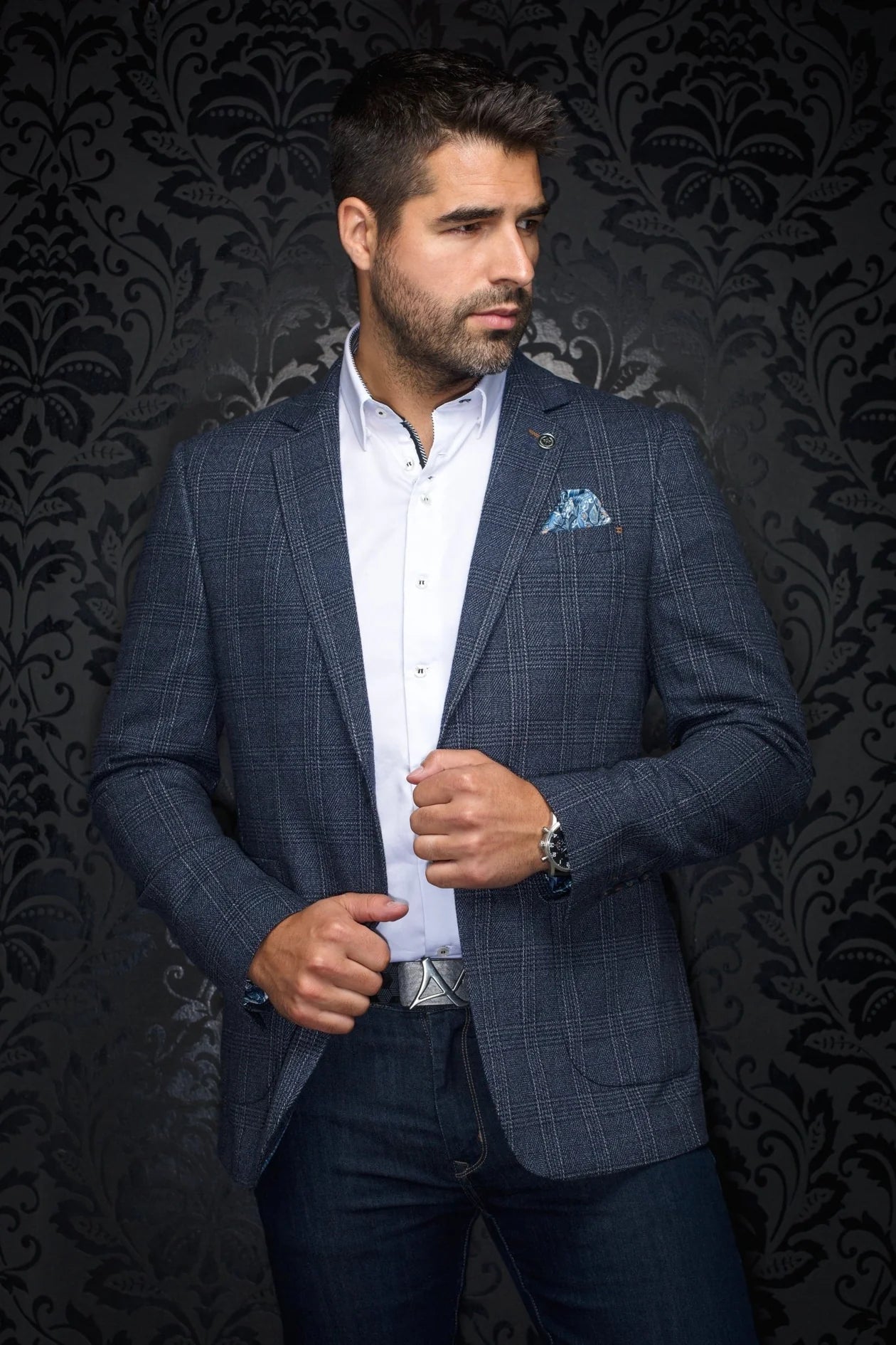 Man wearing a blue checkered suit with a white shirt and patterned pocket square against a dark, decorative wall.