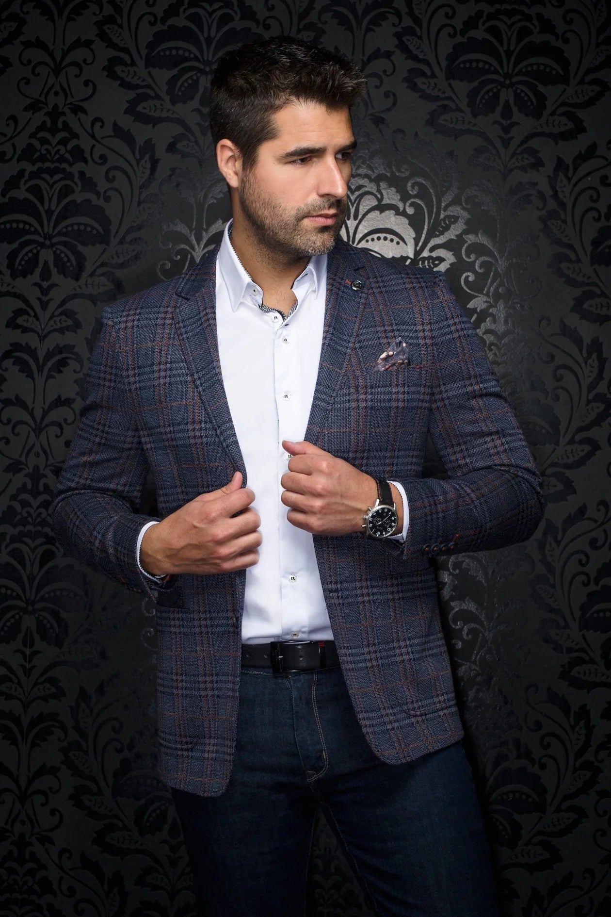 Man wearing a patterned blazer over a white shirt against a dark, ornate wall.