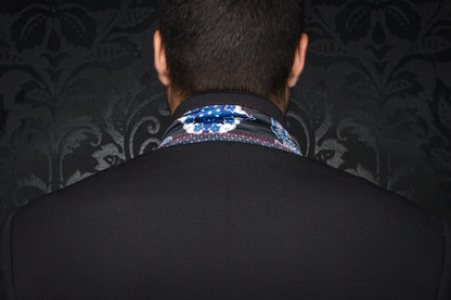 Person wearing a dark suit with a patterned shirt collar against a dark, textured wall.