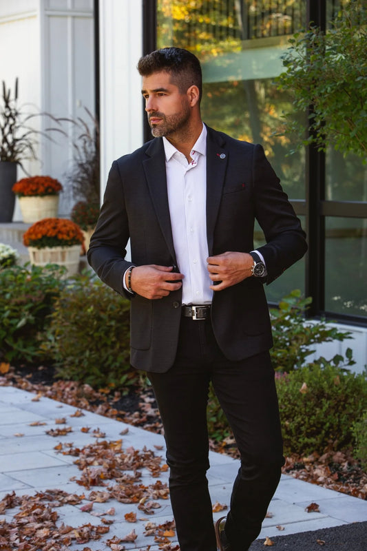 Man in a black suit standing outdoors with plants and a building in the background
