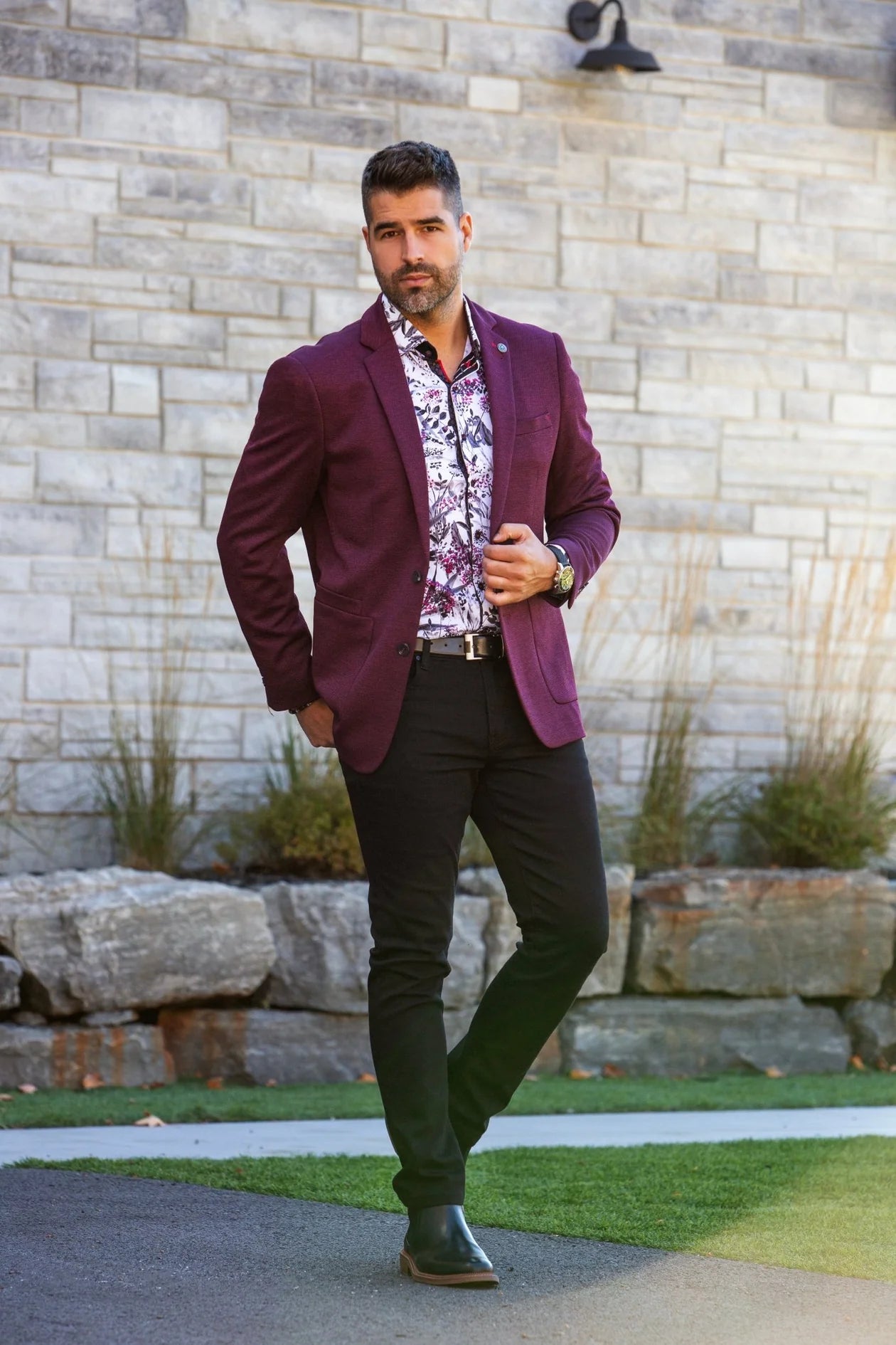 Man wearing a purple blazer over a patterned shirt with green pants against a stone wall.