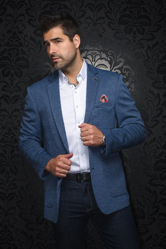 Man wearing a blue blazer over a white shirt with a patterned pocket square against a dark, ornate wall.