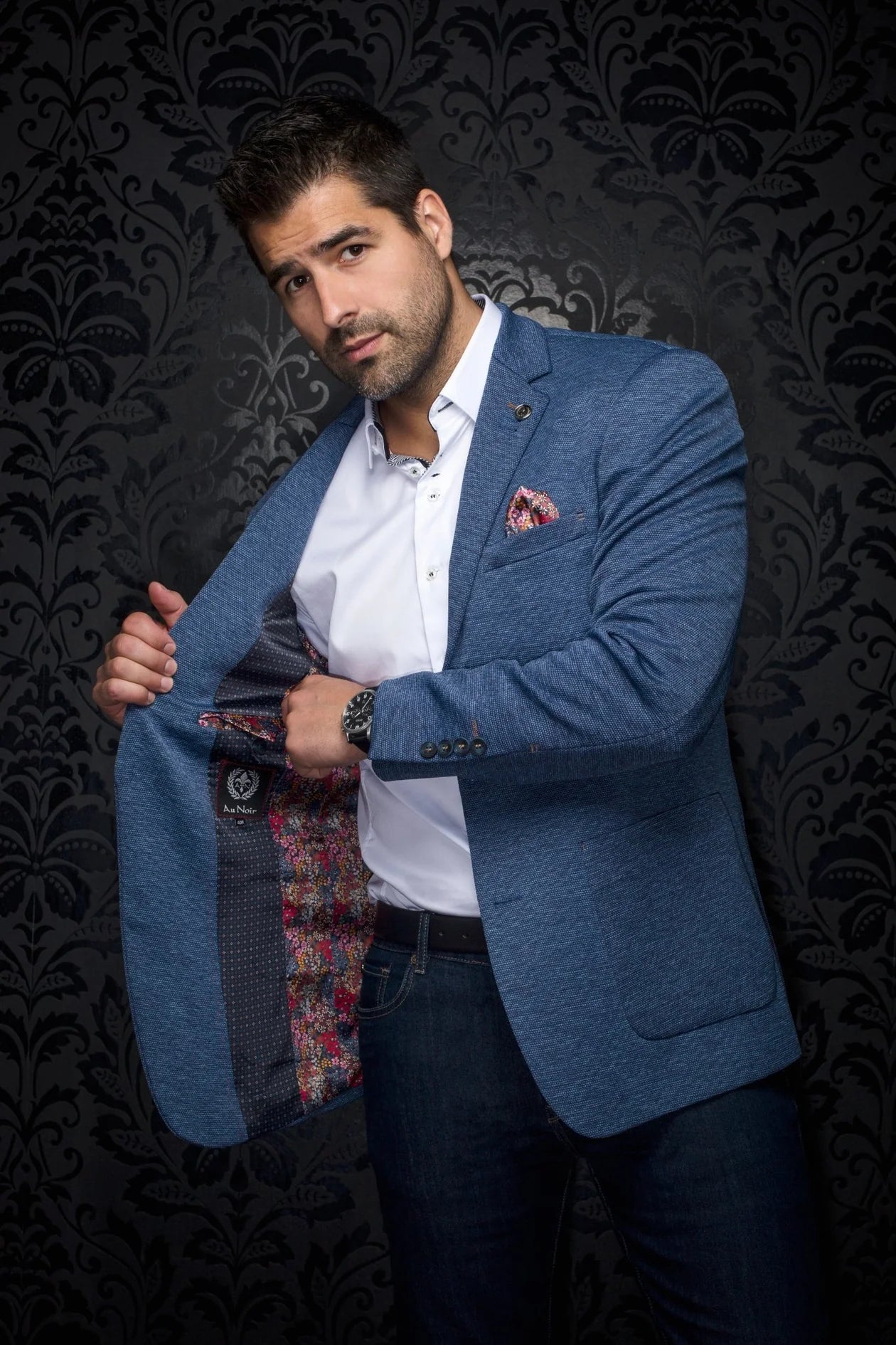 Man wearing a blue blazer over a white shirt with a patterned tie against a dark, decorative background