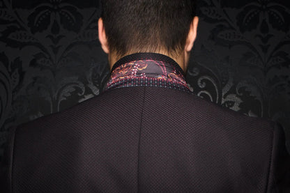 Back view of a person wearing a dark suit with a patterned shirt collar against a dark, ornate wall.