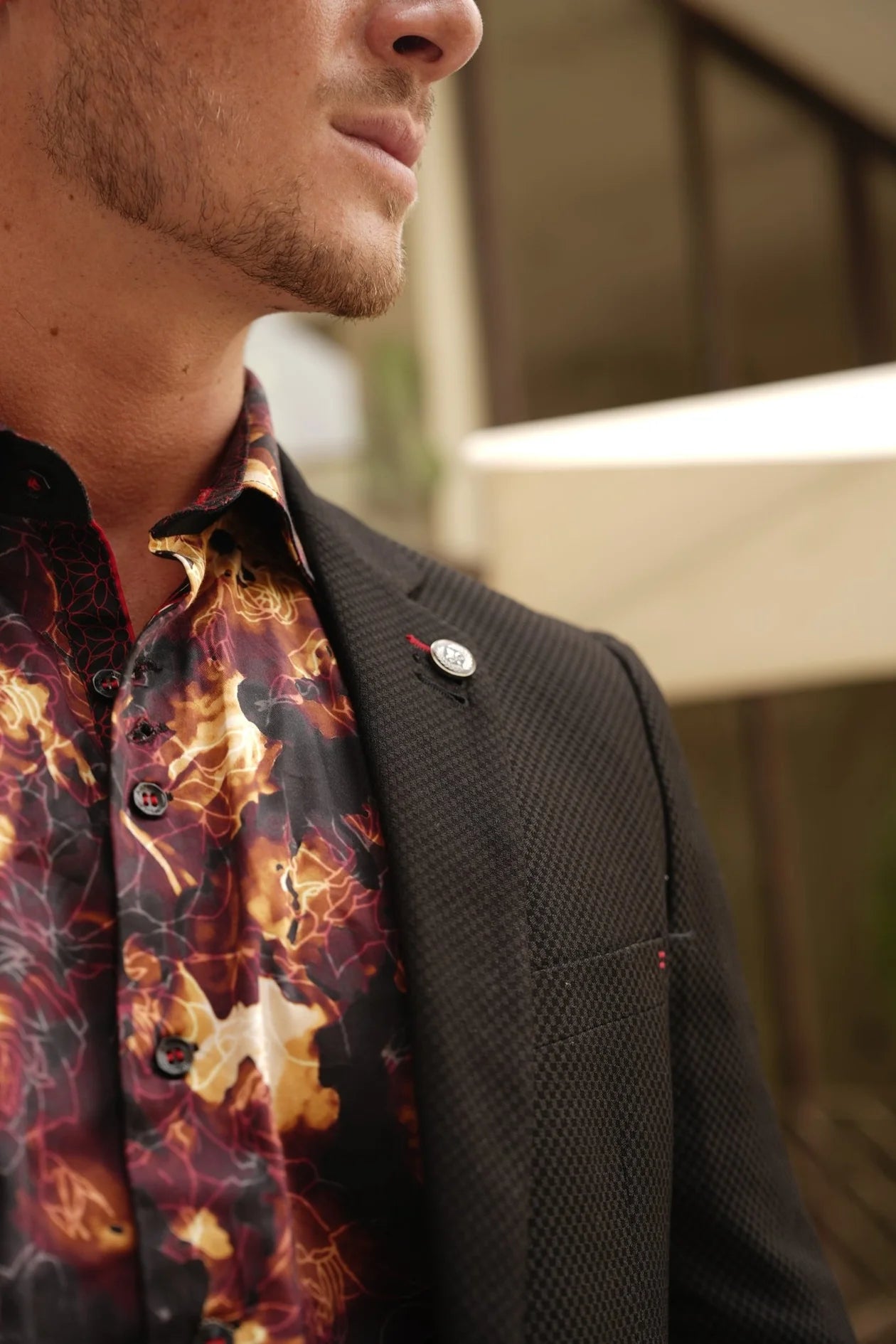 Man wearing a dark jacket over a fiery patterned shirt with a blurred background