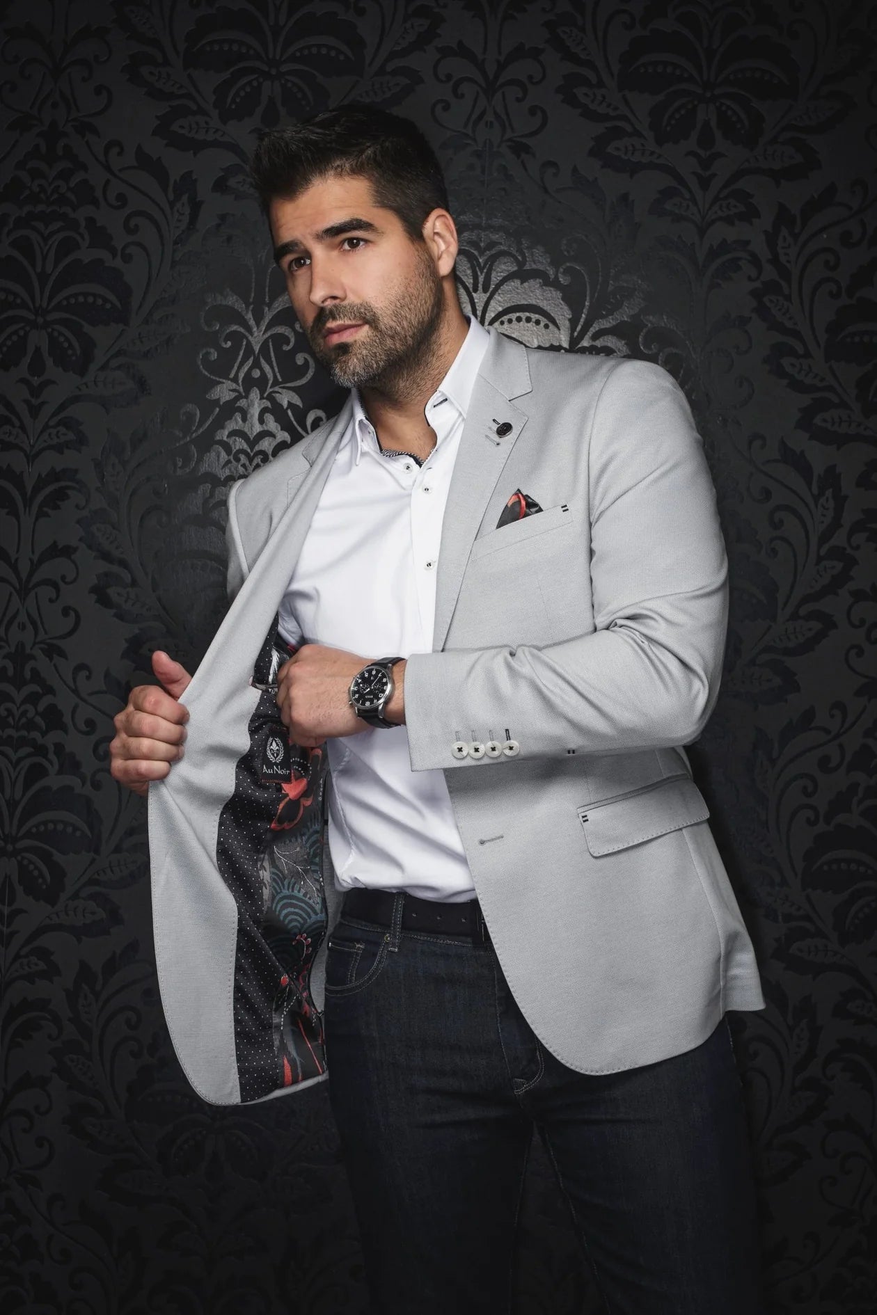 Man in a light gray blazer over a white shirt with a patterned tie against a dark, ornate wall.