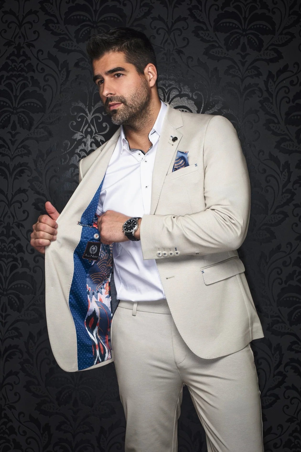 Man in a light-colored suit with a patterned shirt against a dark, decorative wall.