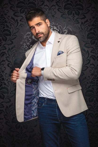 Man wearing a beige blazer over a white shirt with a patterned tie against a dark, ornate wall.