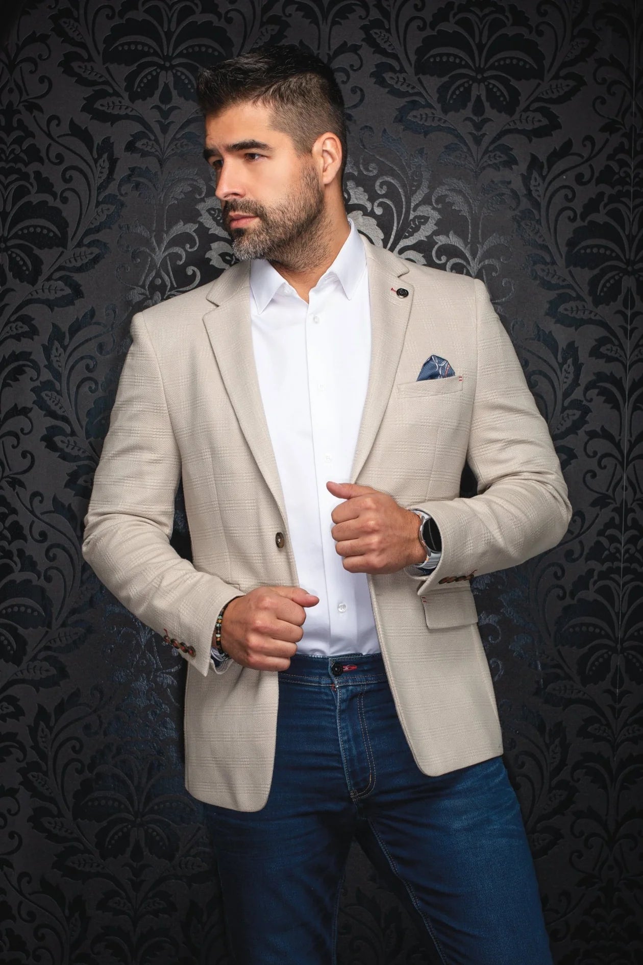Man wearing a beige blazer over a white shirt with blue jeans against a dark patterned wall.