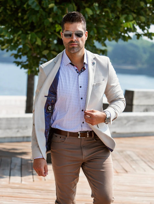 Man in a light-colored blazer and patterned shirt standing outdoors by a body of water.