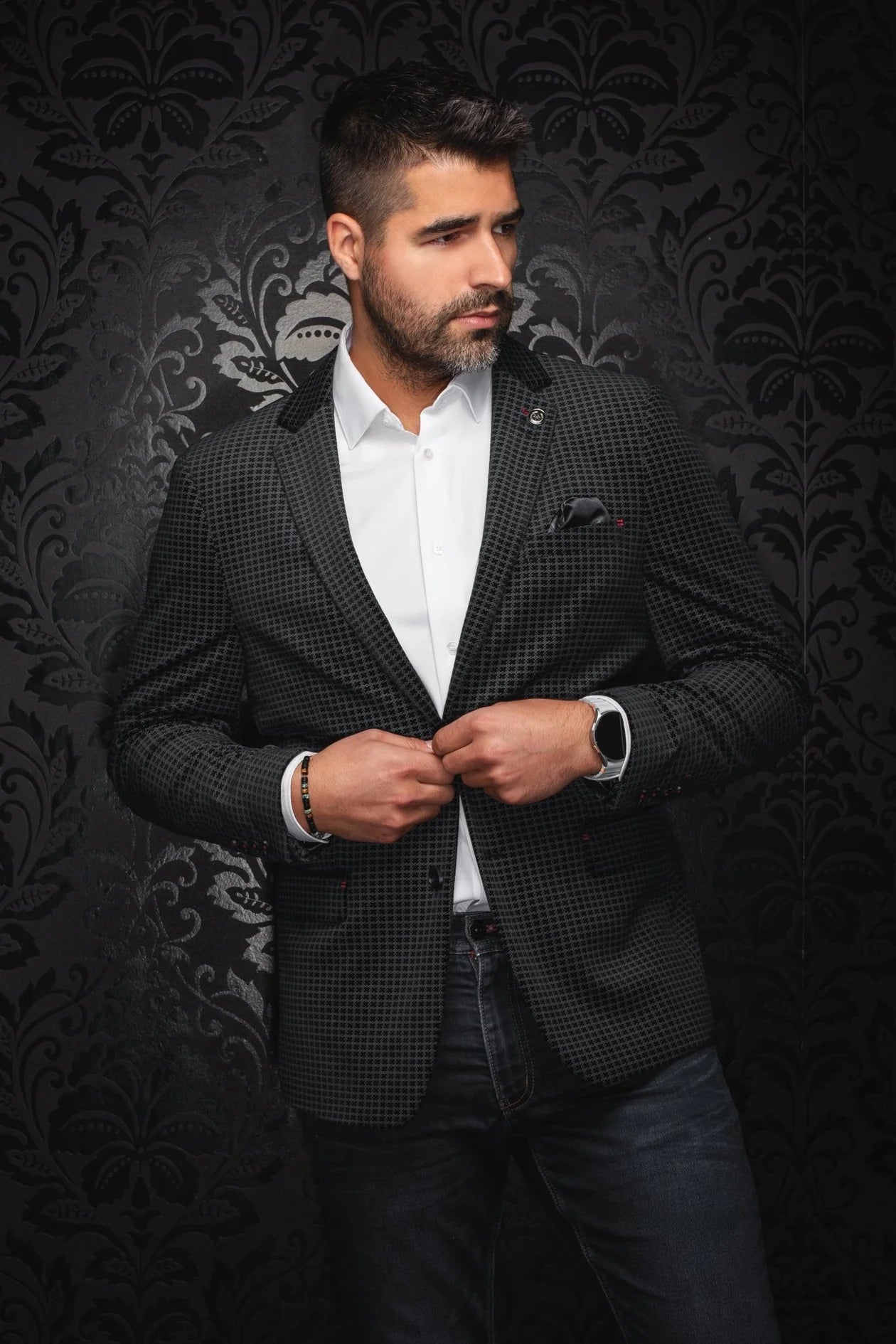 Man wearing a black checkered blazer over a white shirt against a dark patterned background