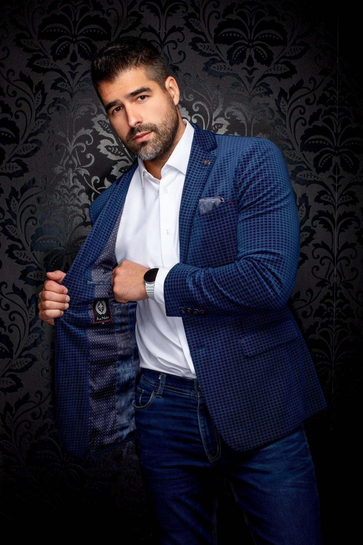 Man wearing a blue checkered blazer over a white shirt with a dark patterned background