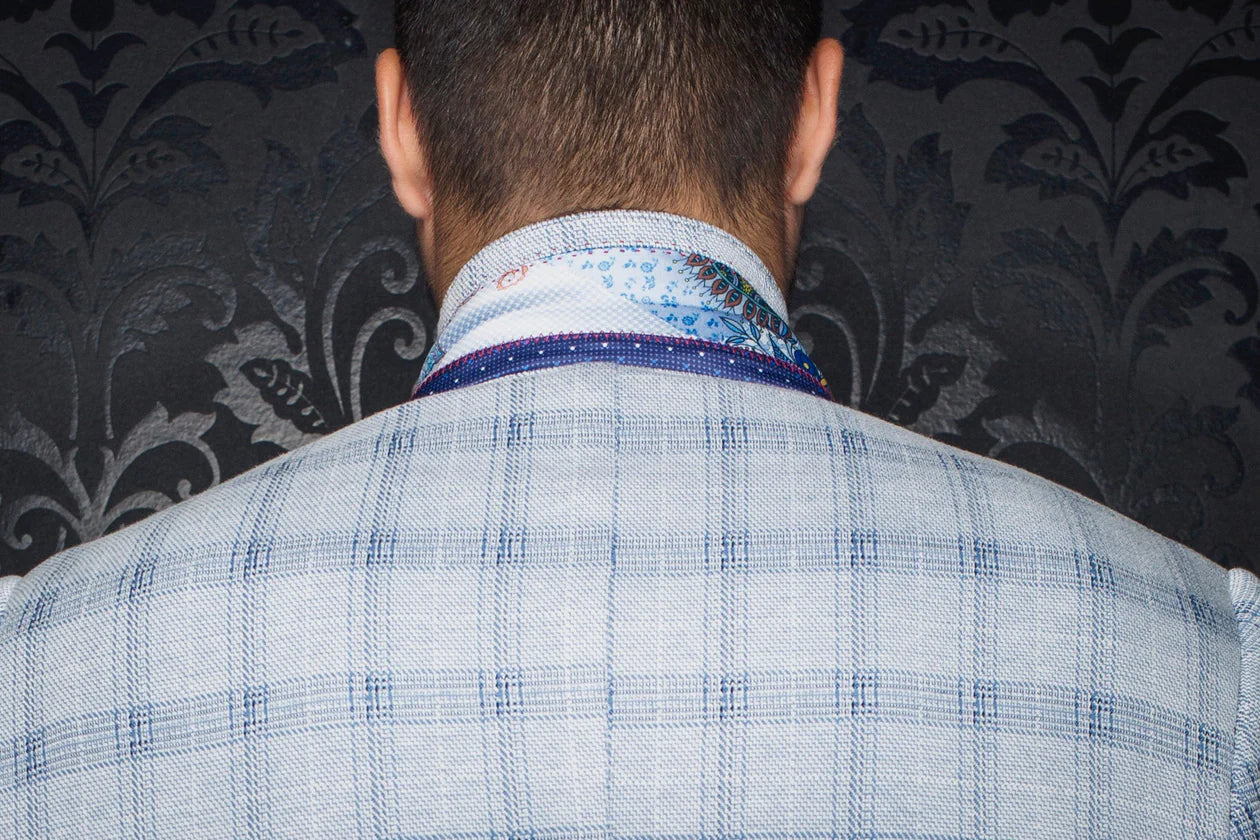 Back view of a person wearing a blue checkered suit jacket with a patterned shirt underneath against a dark floral wallpaper.