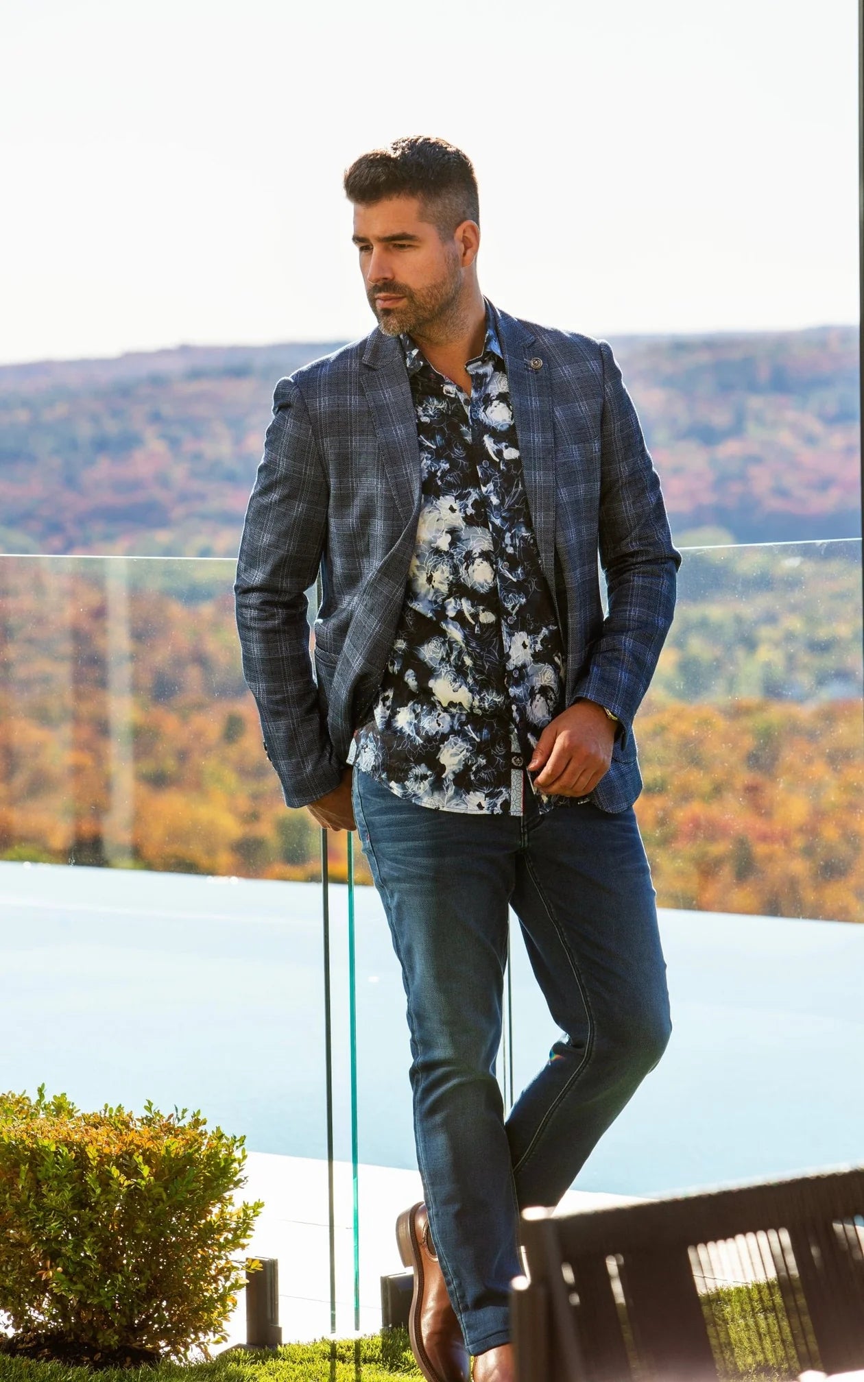 Man in a stylish outfit with a jacket, floral shirt, and jeans standing outdoors with mountains in the background.