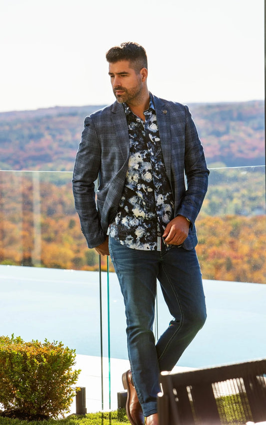 Man in a stylish outfit with a jacket, floral shirt, and jeans standing outdoors with mountains in the background.