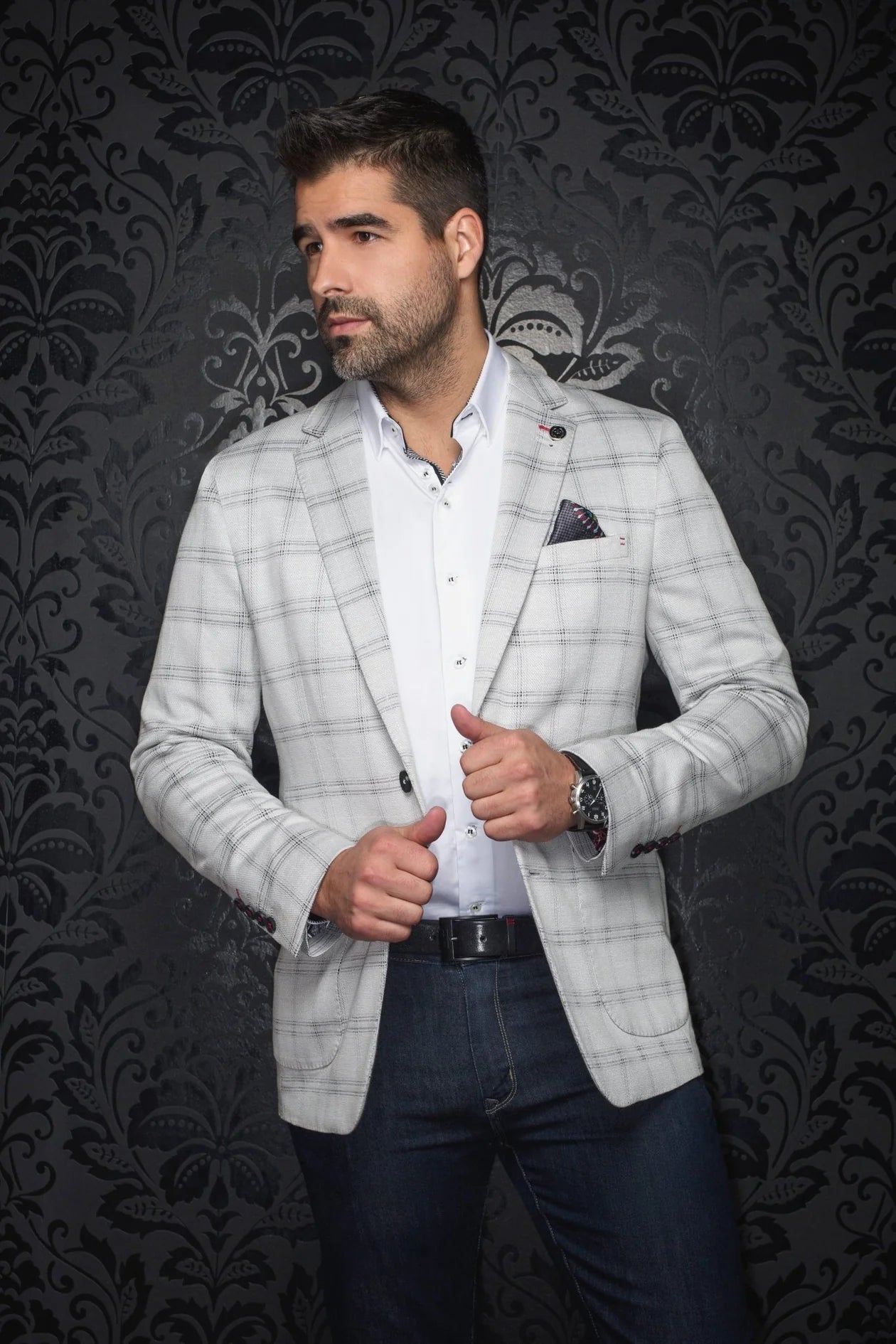 Man wearing a light gray checkered blazer over a white shirt with a dark patterned wall background