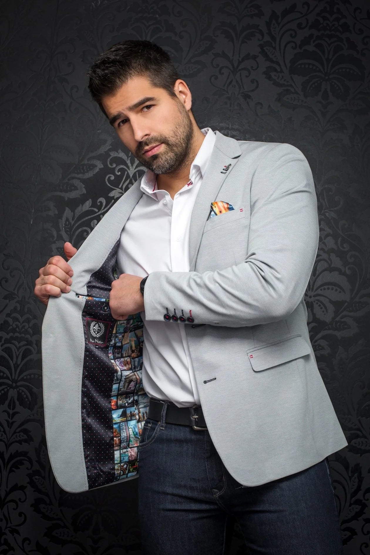 Man wearing a light gray blazer over a white shirt with a patterned pocket square, against a dark decorative background.