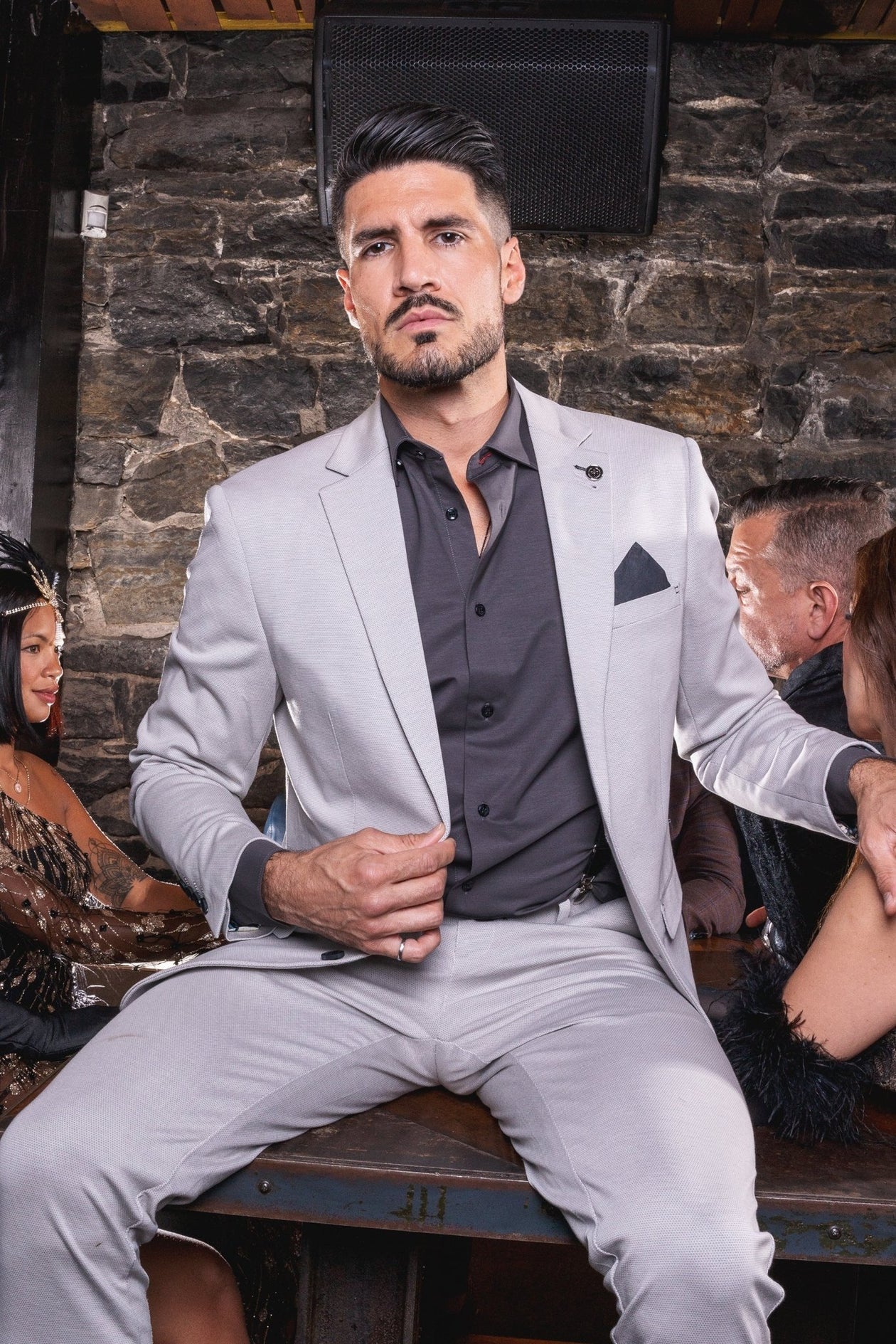 Man in a gray suit sitting in a social setting with stone wall background