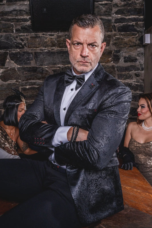 Man in a formal suit standing in a social setting with people in the background.